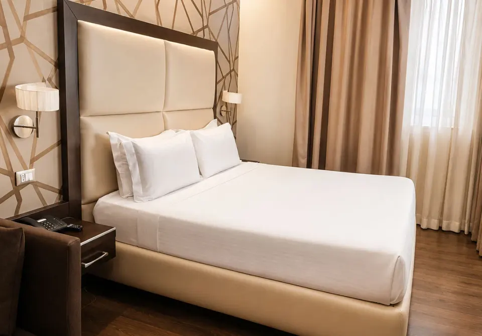A double bed with white linen and a tall padded headboard against a wall with a gold geometric pattern, lit by a wall lamp.
