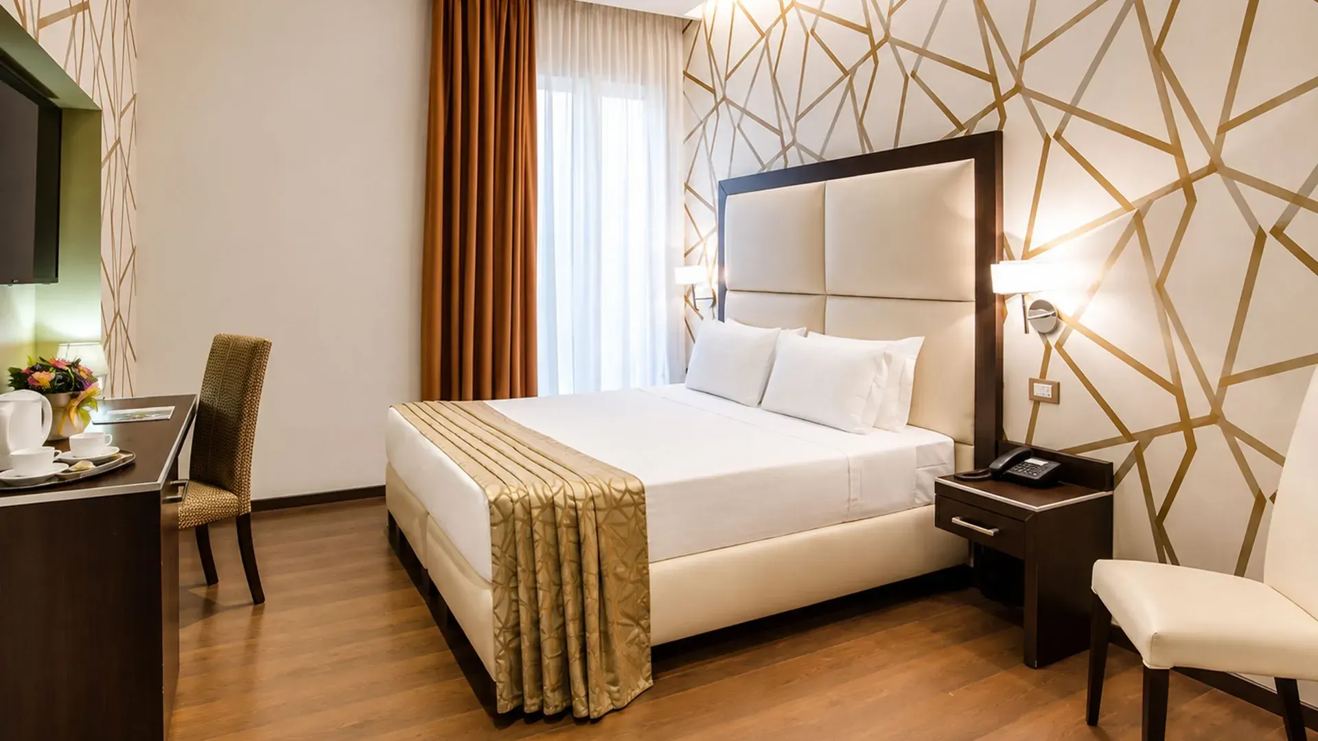 A bedroom with a cream padded headboard, a gold geometric pattern on the wall, rust-coloured curtains and a gold runner across the bed.